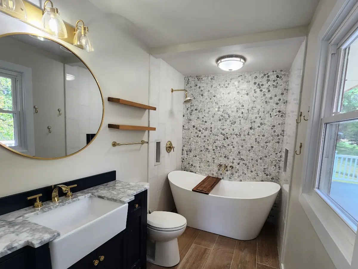 bathroom remodel 1 after, modern bathroom, white bathtub, big round golden framed mirror, black bathroom cabinets with white and gray marble countertop, golden fixtures and wooden floor